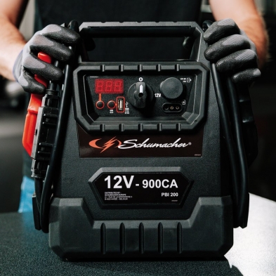 Booster Schumacher 12V 900CA en uso, con empuñadura ergonómica y cables de arranque profesionales.