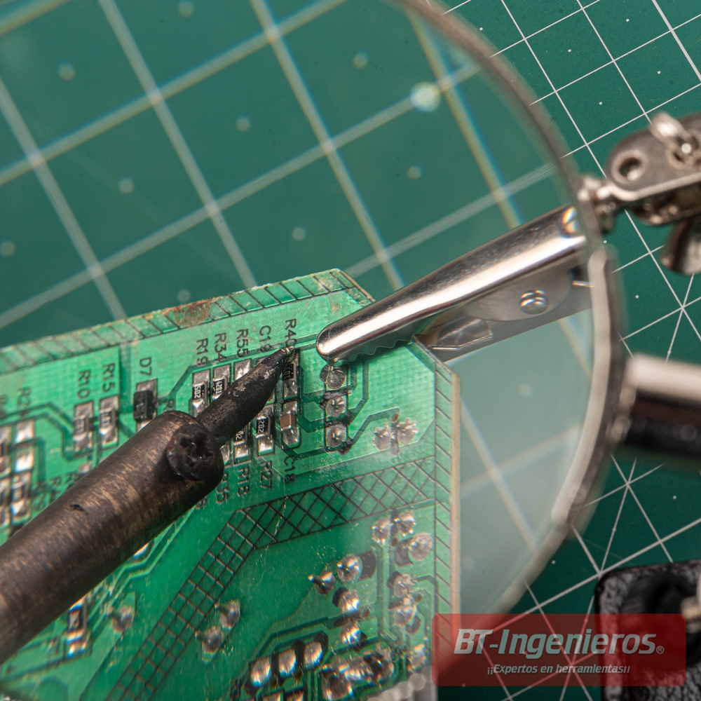 Tercera mano con lupa sujetando una placa electrónica mientras se realiza una soldadura de precisión